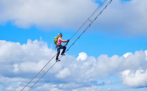 Frau klettert auf dem Donnerkogel Klettersteig über eine Hängebrücke, umgeben von blauem Himmel und Wolken.