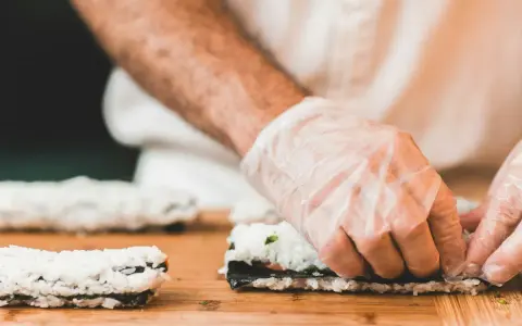 Hände mit Handschuhen rollen Sushi auf einem Holzbrett. So einfach ist Sushi selbst machen!