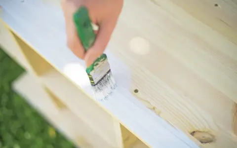 Pędzel nakłada białą farbę na surowe drewno sosnowe, tworząc gładką powierzchnię. Idealne rozwiązanie, czym pomalować surowe drewno sosnowe.