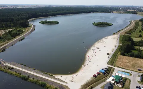 Letnie słońce nad jeziorem opolskim. Ludzie odpoczywają na piaszczystej plaży, pływają w wodzie. W oddali las i zabudowania.