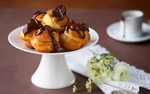 Un plateau blanc présente des profiteroles gastronomiques garnies de crème et nappées de chocolat noir. Des fleurs délicates ajoutent une touche de fraîcheur.