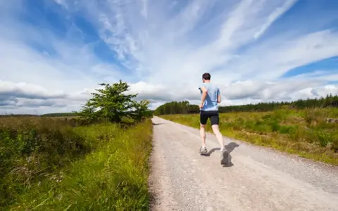 Uomo corre su strada sterrata, ideale per la preparazione 10 km principianti. Cielo azzurro con nuvole bianche.
