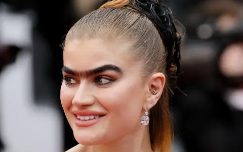 Primer plano de una mujer con una uniceja prominente, cabello recogido en coleta y pendientes llamativos.