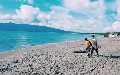 Due ragazzi passeggiano sulla spiaggia della Maremma Toscana, uno con una tavola da surf, mentre il mare azzurro lambisce la riva.