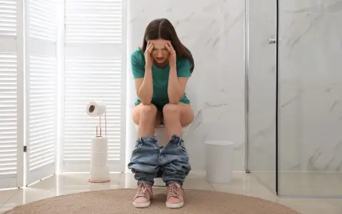 Donna seduta sul water con le mani sulla testa, visibilmente sofferente. Sembra che abbia mangiato troppo e ora si senta male, come se avesse "feci a palline".
