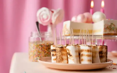 Rostade marshmallows på spett, perfekt för roliga recept att baka till kalaset. En burk strössel och en tårta i bakgrunden.