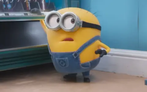 Minionek w ogrodniczkach zastanawia się, po jakim języku mówią minionki, gdy patrzy na automat z przekąskami.