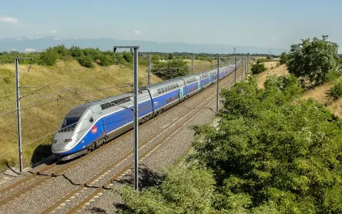 Niebiesko-srebrny pociąg dużych prędkości TGV na torach kolejowych, otoczony zielenią i słupami trakcyjnymi.