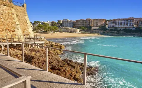 Paseo marítimo de madera junto a rocas y mar turquesa, con playa y edificios al fondo. Un tramo del camí de ronda Tarragona.