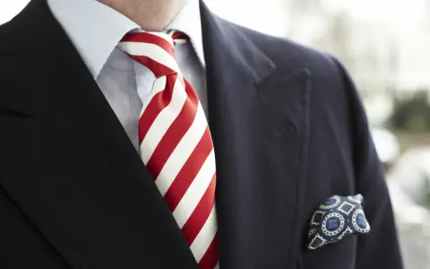 Elegancki mężczyzna w granatowym garniturze, błękitnej koszuli i z wyrazistym, czerwonym krawatem w paski. W kieszeni marynarki chusteczka w niebieskie wzory.