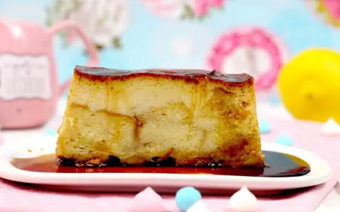 Un trozo de pan de calatrava, con su caramelo dorado, reposa en un plato blanco. Al fondo, una taza rosa y un limón.