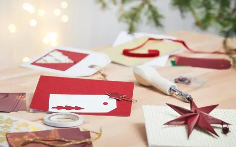 Förberedelser för att måla julkort. Rött glitterpapper, vita etiketter med julgransmotiv och en stjärnformad gemklämma ligger på ett träbord.