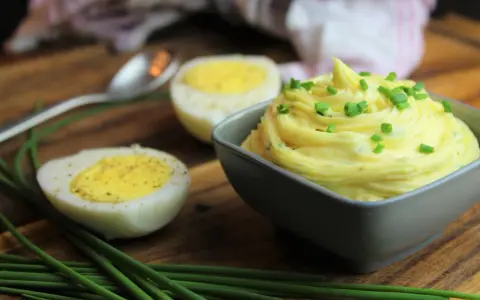 Cremige Sauce Tartar, garniert mit Schnittlauch, neben halbierten Eiern auf einem Holzbrett.