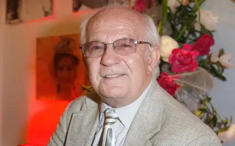 Uśmiechnięty Jerzy Antczak, reżyser znany z niezapomnianych filmów, w eleganckim garniturze, na tle bukietu kwiatów i portretu.
