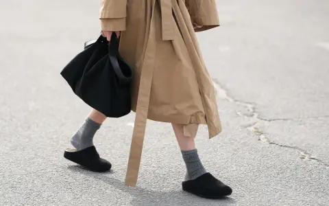 Persona camina con zuecos negros y calcetines grises, llevando un bolso negro y un abrigo beige.