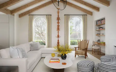 Salón luminoso con vigas de madera en el techo, creando la sensación de casas con techos bajos. Sofá blanco, mesa ovalada y butaca a rayas.