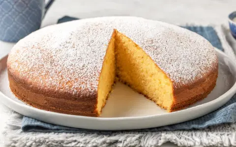 Torta soffice allo yogurt greco, cosparsa di zucchero a velo, con una fetta tagliata che ne rivela l'interno dorato.