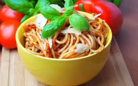 Pyszne spaghetti bez mięsa w żółtej misce, udekorowane bazylią i parmezanem. Obok pomidory i papryka.