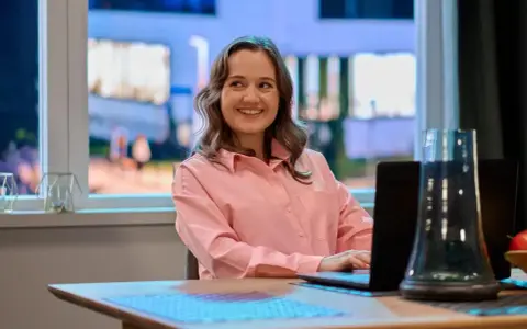 Uśmiechnięta Dorota Krempa pracuje przy laptopie, siedząc przy stole. W tle widać okno z nocnym widokiem.