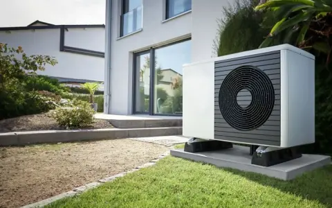 Un moderno sistema hvac, una unidad exterior blanca con rejilla circular negra, se asienta sobre una base de hormigón en un jardín cuidado.