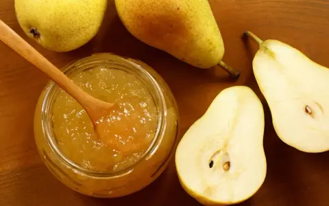 Un pot de confiture de poires maison, avec une cuillère en bois, entouré de poires fraîches coupées et entières sur fond de bois.