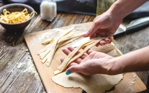 Hände formen frische Nudeln auf einem Holzbrett. Teigstreifen liegen bereit, daneben eine Schüssel mit fertigen Nudeln.