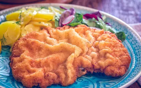 Ein knuspriges, originales Wiener Schnitzel auf einem blauen Teller, serviert mit Kartoffelsalat und frischem Grün.
