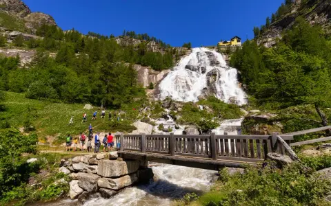 Cascata imponente con escursionisti su un sentiero, un ponte di legno attraversa un torrente. Un esempio di cosa vedere in Piemonte per gli amanti della natura.