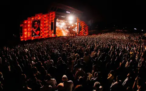 Un enorme pubblico al festival in Portogallo, illuminato da un palco con luci arancioni e un'immagine su schermo.