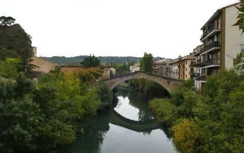 El puente de la cárcel de Estella cruza un río sereno, reflejando su arco de piedra entre frondosa vegetación y edificios históricos.