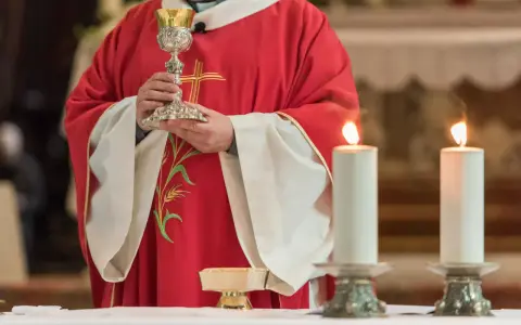 Kapłan w czerwonej szacie z krzyżem trzyma kielich podczas konsekracji. Płoną dwie świece.