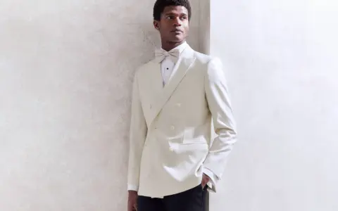 A Black tie wedding guest man in a cream double-breasted tuxedo with a matching bow tie, looking dapper.