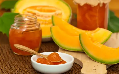Marmellata di melone in vasetti, con fette di melone fresco e un cucchiaino pieno di confettura.