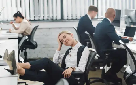 Zmęczona pracownica z nogami na biurku, jakby zastanawiała się, czy urlop przechodzi do nowej pracy. W tle inni pracują.