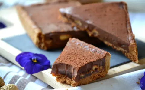 Tranche de tarte façon snickers, garnie de chocolat et de cacahuètes, saupoudrée de cacao. Une fleur bleue décore la présentation.
