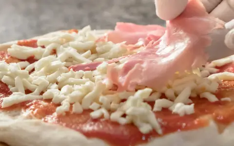 Ręka w białej rękawiczce kładzie plasterki szynki na pizzy z serem i sosem pomidorowym.