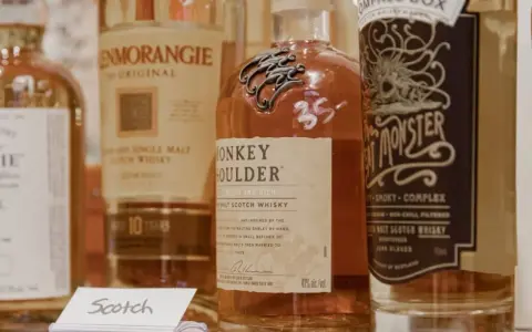 Flera flaskor whisky, inklusive Monkey Shoulder och Glenmorangie, står uppradade. En liten skylt med texten "Scotch" syns framför.