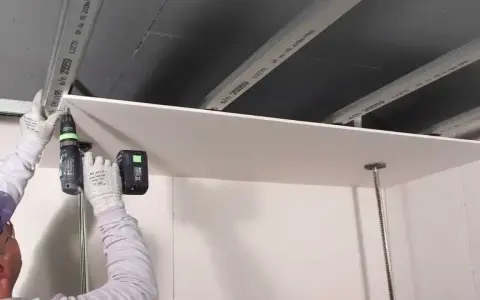 Persona con guanti avvita una lastra di cartongesso al soffitto metallico, un progetto di cartongesso fai da te.