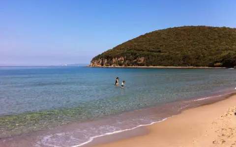 Spiaggia della Maremma con due bambini che giocano nell'acqua cristallina, sotto un cielo azzurro e una collina verdeggiante.