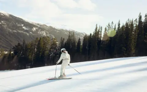En person åker skidor utför en snöig backe, omgiven av granar och snöklädda berg. En perfekt dag på en av Sveriges skidorter.