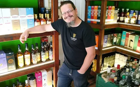 Mann in einem Guinness-T-Shirt steht in einem Laden voller Whisky-Flaschen und -Schachteln. Er lächelt und lehnt sich an ein Regal.