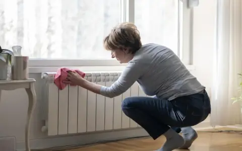 Mujer limpiando un radiador blanco con un paño rosa. Un ejemplo de cómo limpiar radiadores para mantener el hogar cálido y limpio.