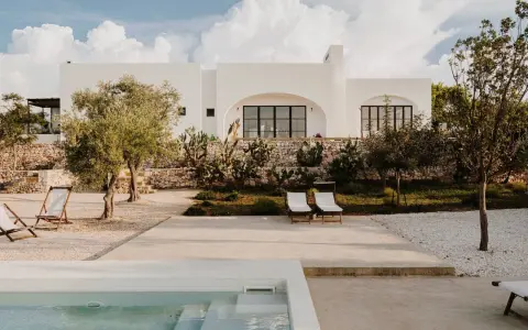 Piscina e lettini in una delle masserie famose puglia, con ulivi e cactus sotto un cielo azzurro con nuvole bianche.