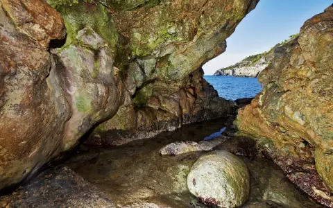 L'entrata di una grotta marina, la cala del bove, con rocce muschiose e un piccolo specchio d'acqua che riflette il blu del mare e il cielo.