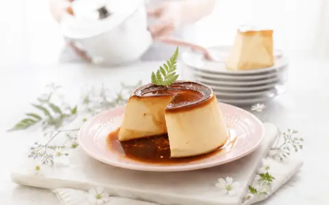 Delicioso flan de queso con caramelo líquido, decorado con una hoja verde. En el fondo, una persona prepara más postres.