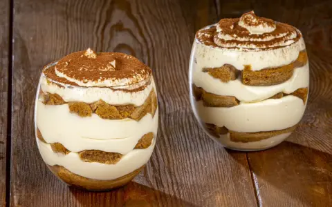 Due bicchieri di tiramisù senza uova, stratificati con crema e savoiardi, spolverati di cacao.