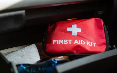 Czerwona apteczka samochodowa z białym krzyżem i napisem "FIRST AID KIT" leży w schowku. Obok niej papierowy ręcznik.