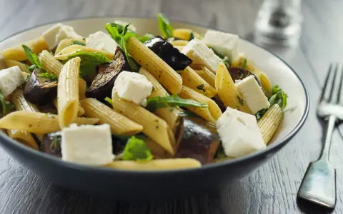 Piatto di pasta con melanzane in bianco, cubetti di formaggio fresco e foglie verdi, servito in una ciotola scura.