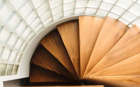 Escalera helicoidal de madera con peldaños que giran bajo un techo de bloques de vidrio translúcido.