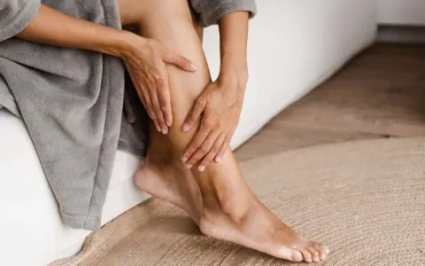 Mani che accarezzano le gambe, evidenziando la pelle secca. La persona è seduta su un letto, avvolta in un accappatoio grigio.